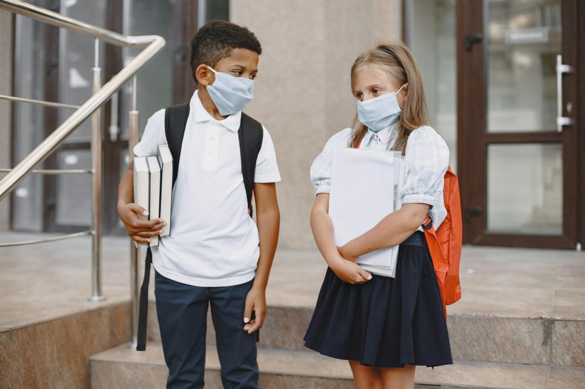Two school children wearing face masks, holding books, standing outside a school building.